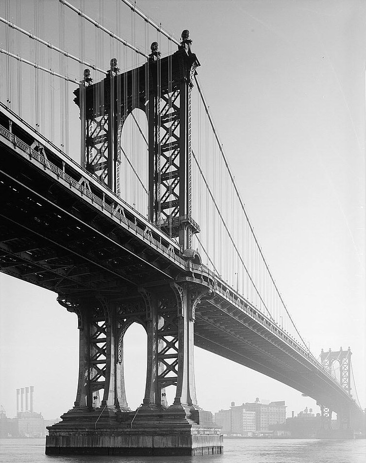 Black and white photograph of the Manhattan Bridge from the water below and to the side, showing the Brooklyn tower, the suspension cables, and the open steel truss soffit of the deck carrying the subway tracks.